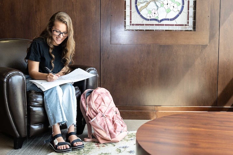 A young woman with long hair and glasses sits on a leather chair, smiling as she writes in a notebook on her lap. A pink backpack rests on the floor beside her in a cozy, wood-paneled room.