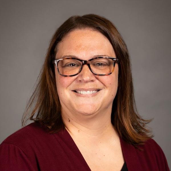 A woman with straight brown hair, glasses, and a beaded necklace smiles at the camera. She is wearing a black top, and the background is plain and gray.