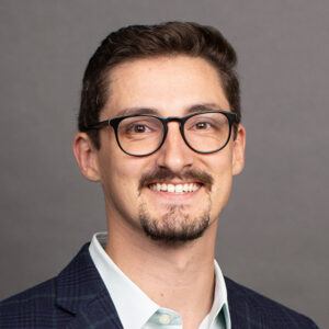 Smiling man with short brown hair, wearing glasses, a trimmed beard, a light shirt, and a dark plaid blazer, posed against a plain gray background.