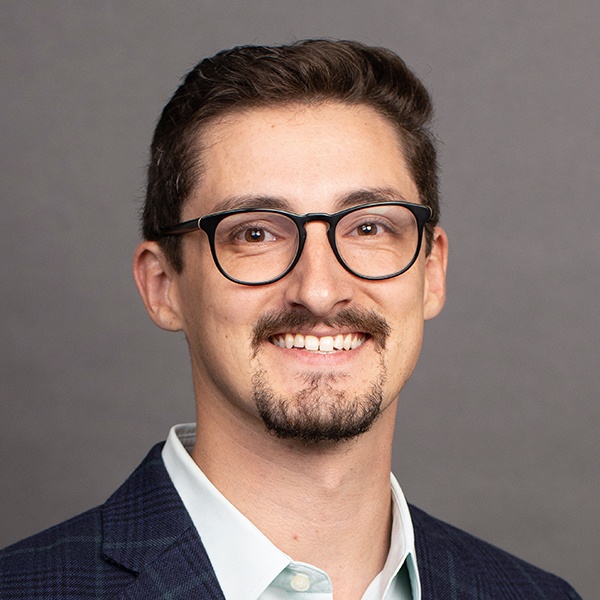 Smiling man with short brown hair, wearing glasses, a trimmed beard, a light shirt, and a dark plaid blazer, posed against a plain gray background.