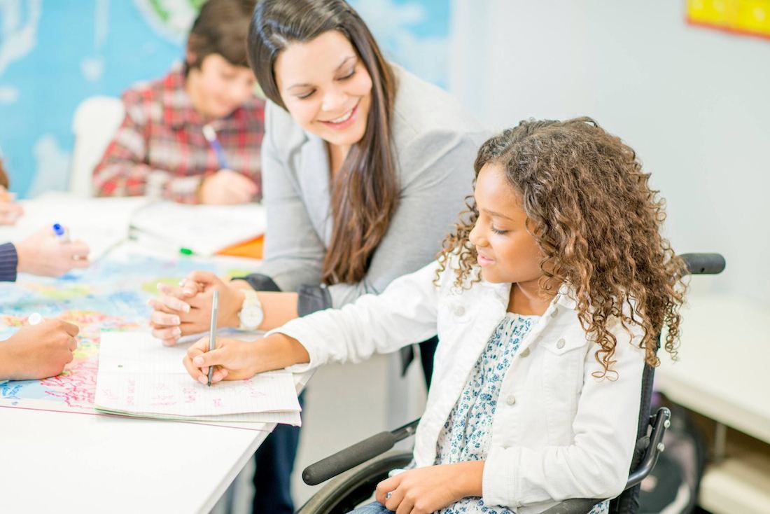 A young girl in a wheelchair writes in a notebook while a smiling teacher watches and assists. Other children are visible in the background, working at a table in a bright classroom.