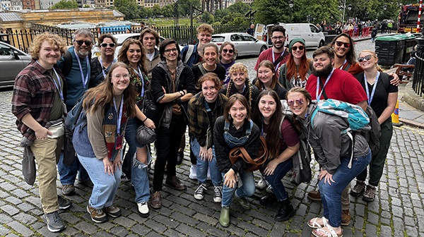 A group of about 20 young adults pose and smile together on a cobblestone street, with trees, buildings, and parked cars visible in the background. They appear happy and are dressed casually.