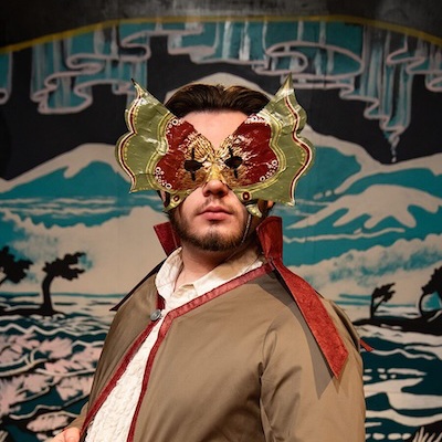 A person wearing a dramatic, butterfly-shaped mask and a costume with a high collar stands in front of a mural featuring mountains, water, and stylized trees.