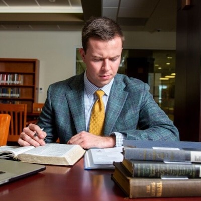 A man in a suit and yellow tie sits at a library table, reading and taking notes with a pen. Several books and a laptop are on the table in front of him. Bookshelves are visible in the background.