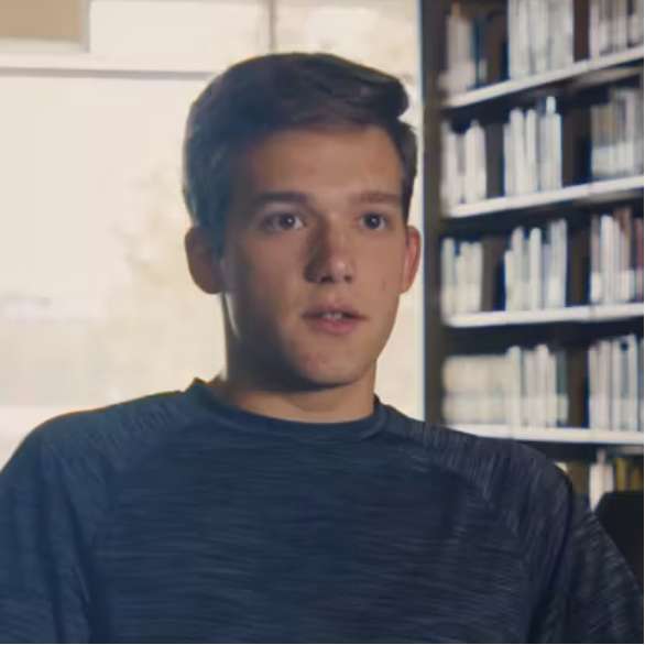 A young man with short brown hair sits indoors near bookshelves, looking slightly to the left. The background shows a window with light streaming in.