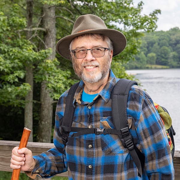 An older man wearing glasses, a brown hat, and a blue plaid shirt stands outdoors by a lake, holding a wooden walking stick and wearing a backpack, with trees and water in the background.