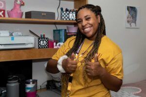 A young woman with long braids smiles and gives two thumbs up. She wears a yellow shirt and a lanyard, with shelves, books, and various items in the background of a dorm or study room.