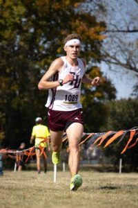 A male cross-country runner in a maroon and white uniform, wearing bib number 1089, runs on a grassy course lined with orange and white ribbons. Trees and another person in a yellow shirt are visible in the background.