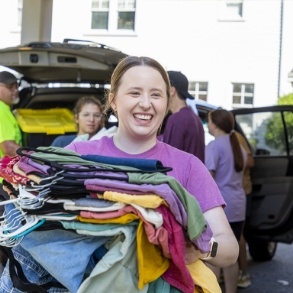 A smiling person holds a large stack of colorful clothes on hangers outdoors, with other people and open car trunks visible in the background.