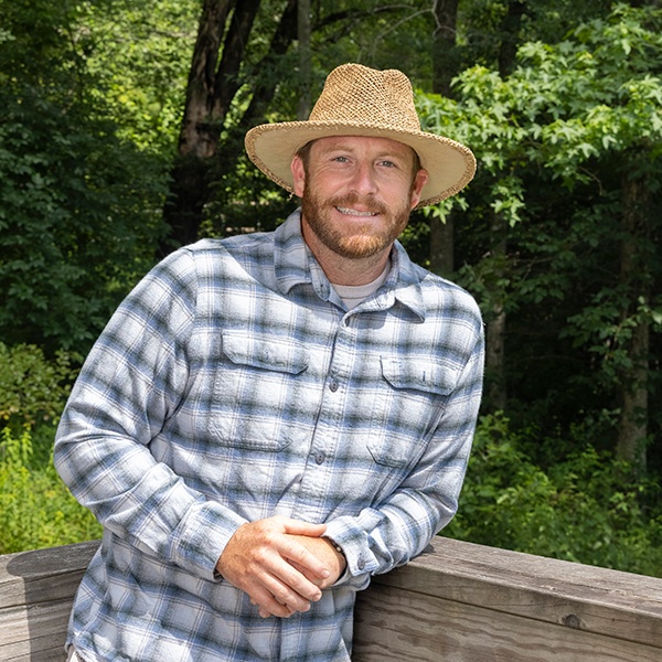 A man wearing a straw hat and a plaid shirt smiles while leaning on a wooden railing outdoors, with green trees and sunlight in the background.