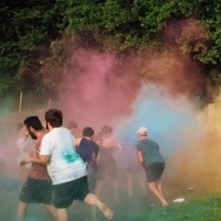 People running and playing outdoors in a cloud of colorful powder, likely at a color run or festival, with trees and grass in the background.