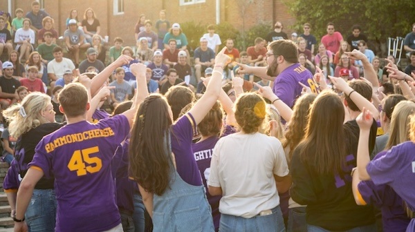 A large group of students wearing purple shirts gathers outside, raising their arms and cheering, with more people seated in the background watching the lively scene.