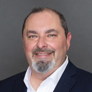 A middle-aged man with short dark hair, a trimmed gray beard, and mustache is smiling. He is wearing a dark suit jacket over a white shirt, set against a plain gray background.