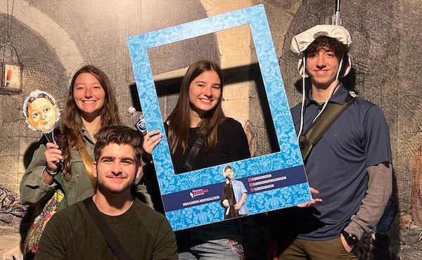 Four young adults pose in a historical-themed setting. One holds a small character face cutout, another holds a large blue picture frame prop, and one wears a historical-style cap. All are smiling.