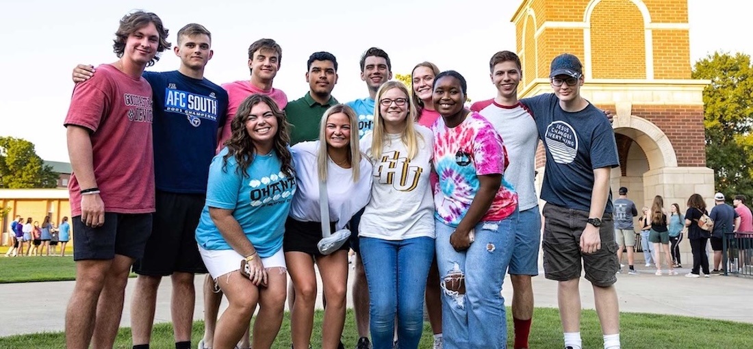 A group of thirteen smiling young adults pose together outdoors in casual clothing, with some wearing college or university shirts, in front of a brick structure on a sunny day.