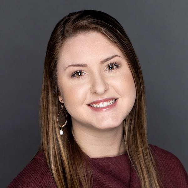 A woman with straight, light brown hair smiles at the camera. She is wearing a maroon top and a dangling earring with a white stone, posing against a plain gray background.