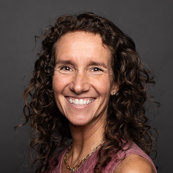 A woman with long, curly brown hair smiles warmly at the camera. She wears a sleeveless mauve top, gold necklace, and small earrings. The background is plain and dark gray.