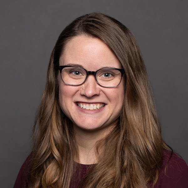 A woman with long brown hair and glasses smiles at the camera in front of a plain gray background. She is wearing a dark-colored top.