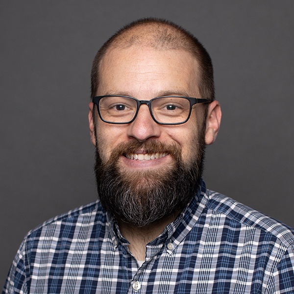 A man with a bald head, full beard, and glasses smiles at the camera. He is wearing a plaid shirt and is posed against a plain, dark gray background.