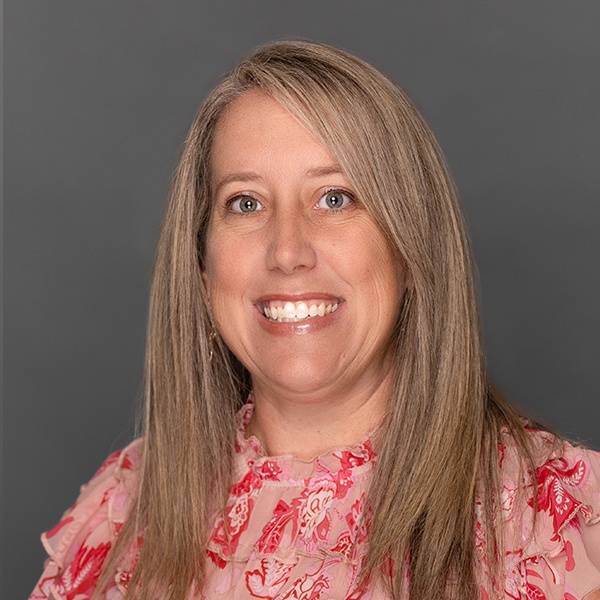 Smiling woman with straight, light brown hair wearing a pink floral blouse, posed in front of a plain gray background.