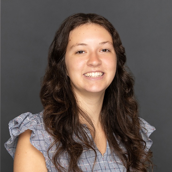A young woman with long, wavy brown hair smiles at the camera. She is wearing a light blue, ruffled short-sleeve top and is posed in front of a plain dark gray background.