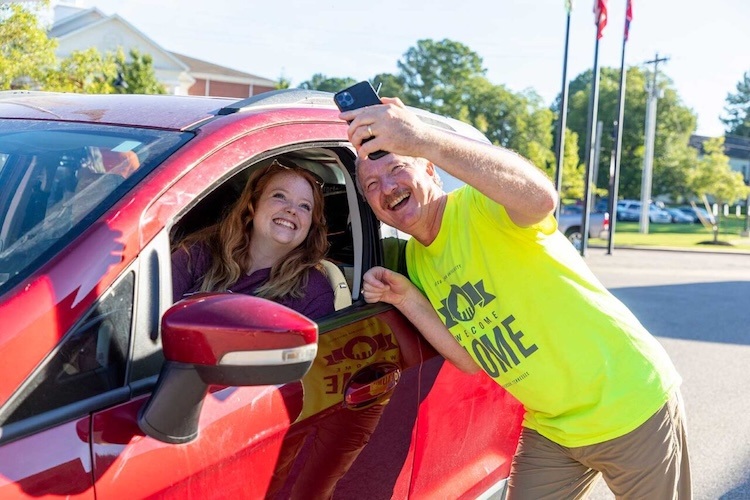 A smiling woman sits in the drivers seat of a red car while a cheerful man in a yellow shirt leans in through the window, taking a selfie of them both outdoors on a sunny day.