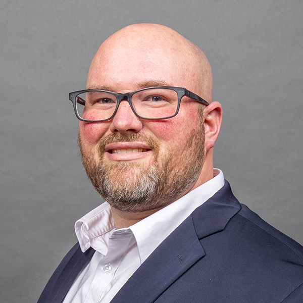A bald man with glasses, a trimmed beard, and a suit jacket over a white collared shirt, smiling in front of a plain gray background.