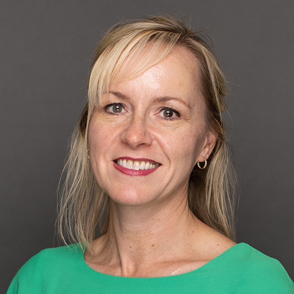Smiling woman with blonde hair wearing a green top and gold hoop earrings, posed against a plain gray background.