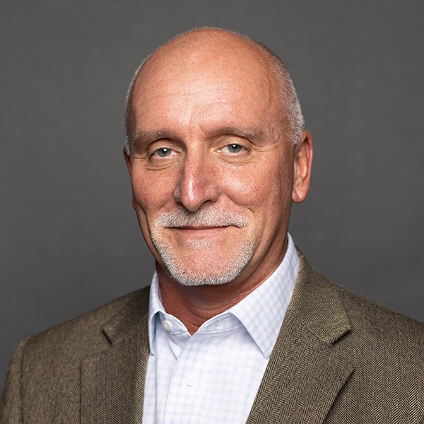 A middle-aged man with a bald head and white goatee, wearing a brown blazer over a white checkered shirt, smiling gently against a plain gray background.