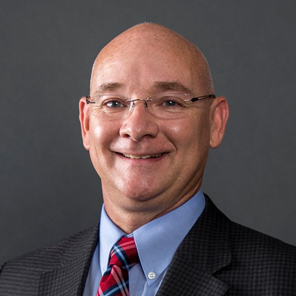 Smiling bald man wearing glasses, a blue shirt, a red and blue striped tie, and a dark checkered suit jacket, posing in front of a plain dark background.
