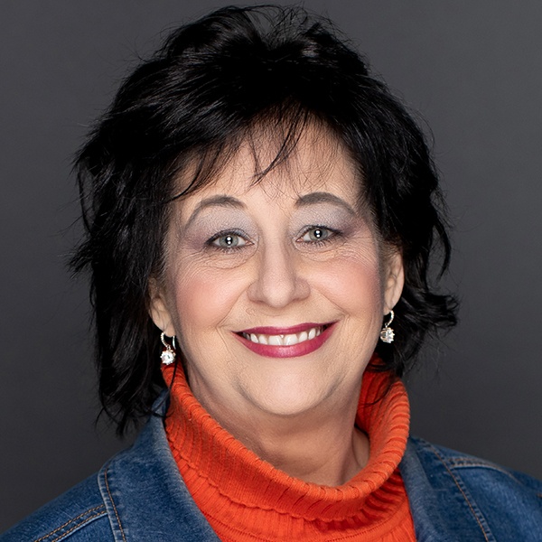A woman with short black hair smiles at the camera. She is wearing a bright orange turtleneck sweater, a denim jacket, dangling earrings, and subtle makeup. The background is a plain dark gray.
