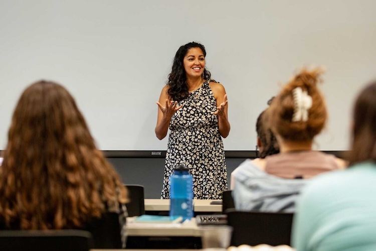 A woman in a floral dress stands smiling and gesturing in front of a classroom, speaking to a group of seated students. The focus is on her, with students’ backs visible in the foreground.