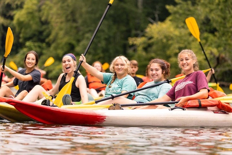 A group of smiling young people paddle in kayaks on a calm river, wearing casual clothes and holding yellow paddles, with green trees in the background.