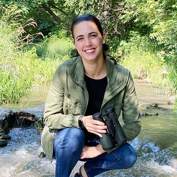 A woman with dark hair in a ponytail, wearing a green jacket and jeans, smiles while crouching by a stream in a lush, green outdoor setting. She holds black binoculars in her hand.