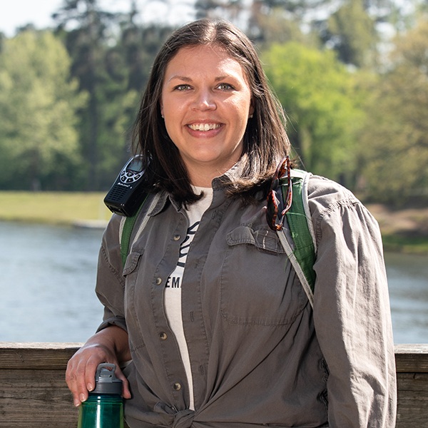 A woman with shoulder-length brown hair smiles at the camera while standing outdoors by a lake. She is wearing a gray shirt, has a walkie-talkie and sunglasses on her shoulder, and holds a green water bottle. Trees are in the background.