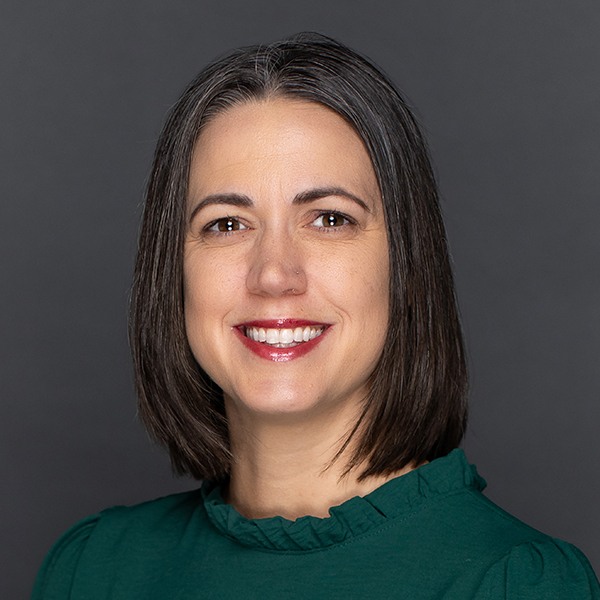 A woman with straight, shoulder-length dark hair with gray streaks smiles at the camera. She is wearing a dark green top with a ruffled neckline and is posed against a plain dark background.