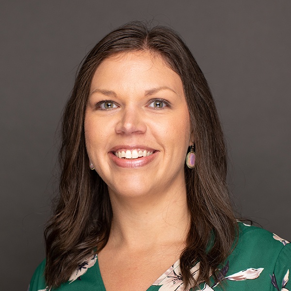 A woman with long brown hair wearing a green floral top and earrings smiles at the camera against a plain dark background.
