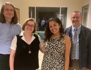 Four adults stand and smile together indoors; two men and two women. One woman wears a black and white dress, another wears glasses and a black vest, while the men wear collared shirts, one with a tie and jacket.