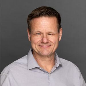 A middle-aged man with short brown hair smiles subtly. He is wearing a light gray collared shirt and is posed in front of a plain gray background.