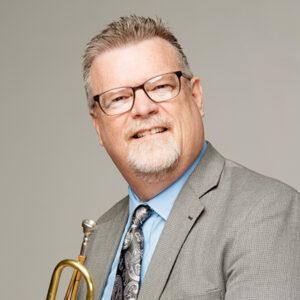 A smiling man with short, light hair and glasses wears a gray suit, blue shirt, and patterned tie. He holds a brass trumpet and poses in front of a plain light background.